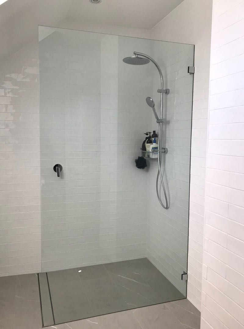 Walk-in frameless fixed panel with polished chrome brackets against white subway tiles and a grey tiled floor